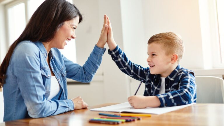 Mother and elementary aged son high-fiving at table while child writes in notebook